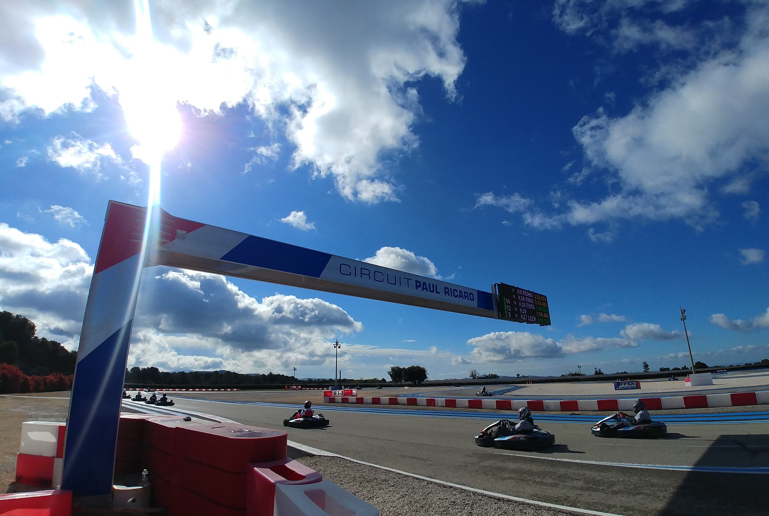 Helmeted kart drivers beneath the Circuit Paul Ricard karting gantry